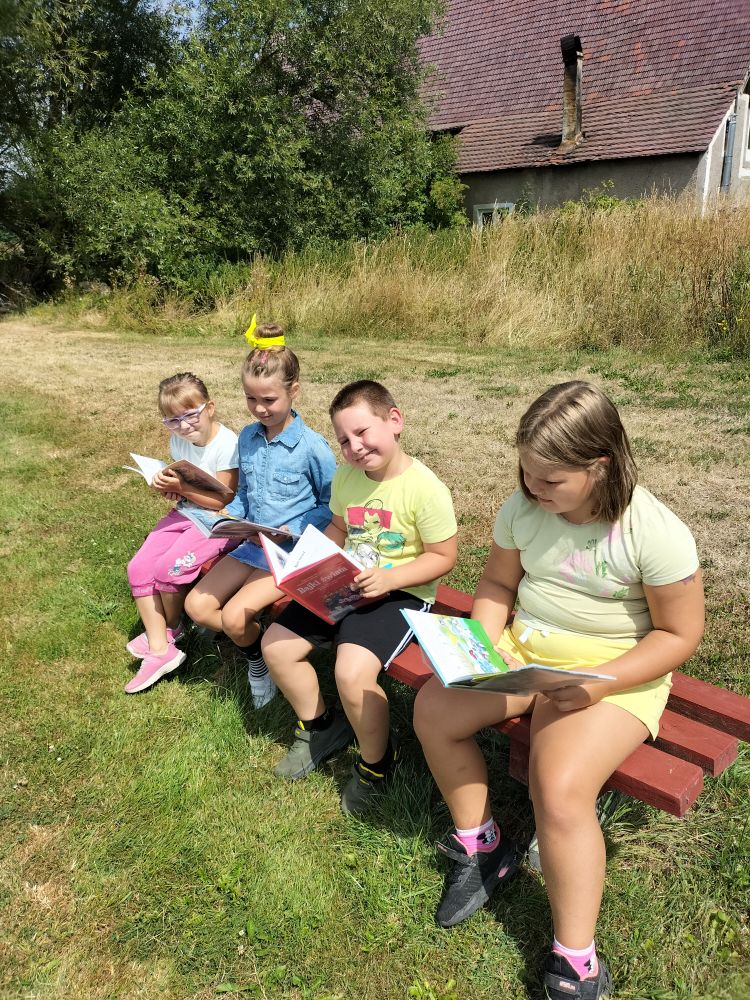 czwórka dzieci siedzi na ławce z książkami w rękach, w tle zielone drzewa, trawa i dom