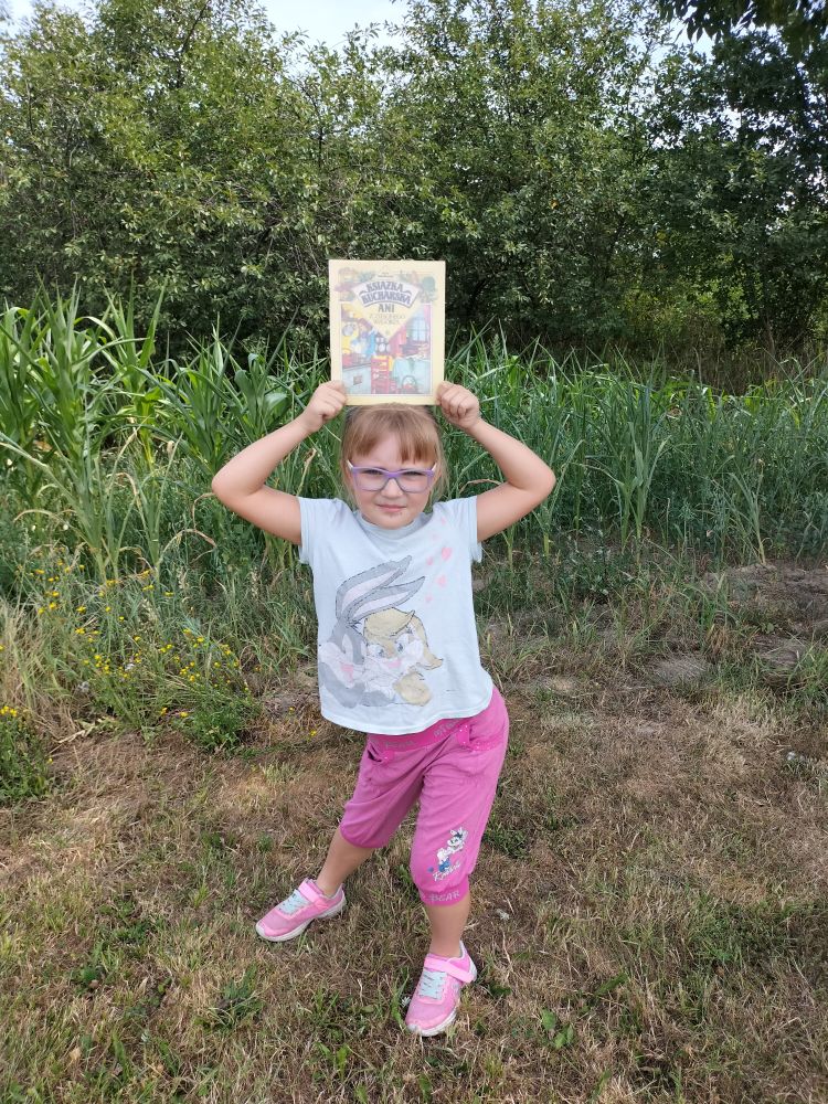 dziewczynka trzyma książkę nad głową, w tle wysoka zielona trawa i drzewa