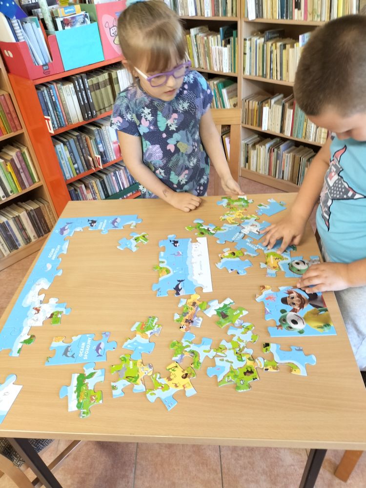 Przy stoliku dziewczynka z chłopcem układają puzzle, w tle regały z książkami