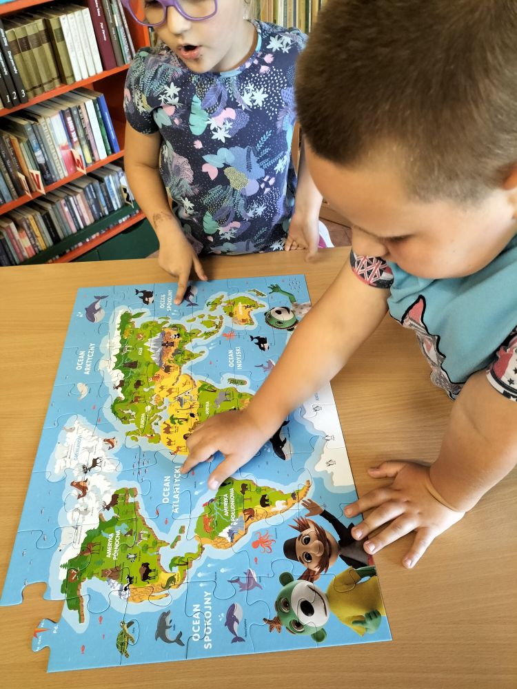 Przy stoliku chłopiec z dziewczynką oglądają kolorową mapę z kontynentami, w tle regały z książkami
