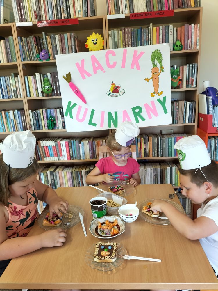 troje dzieci siedzi przy stole w czapeczkach kucharskich i robią słodkie ciasteczka, w tle regały z książkami i plakat – kącik kulinarny