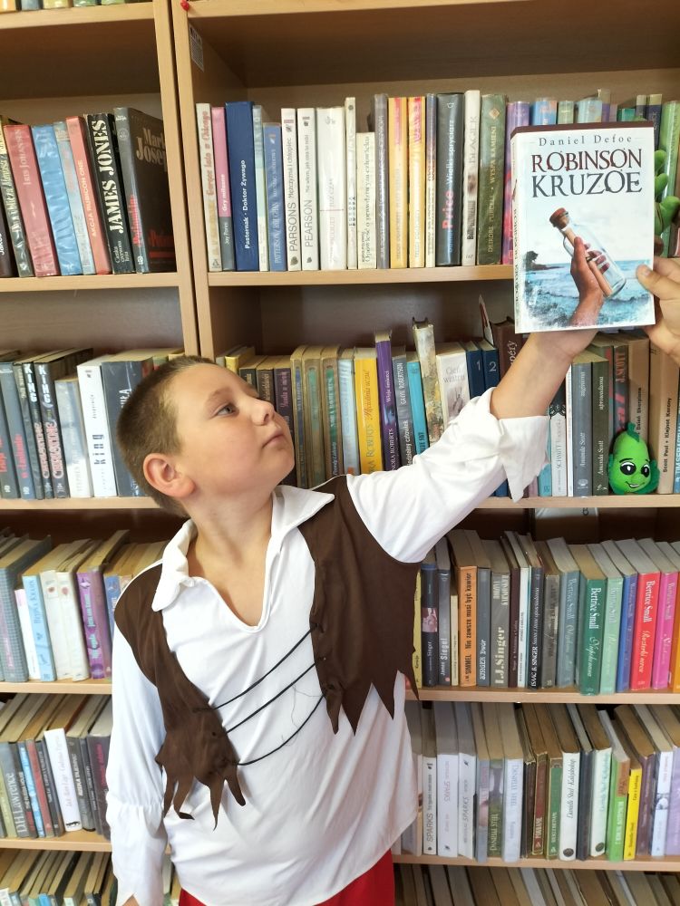 chłopczyk stoi na tle regałów z książkami i trzyma w wyciągniętej, lewej ręce książkę