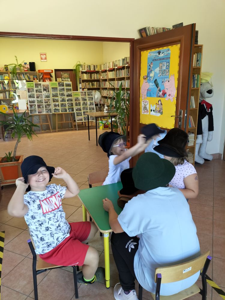 Przy stoliku siedzą cztery osoby w kapeluszach na głowie i biorą udział w zabawie z kapeluszami , w tle regały z książkami, koło fortuny, maskotki i otwarte drzwi biblioteki