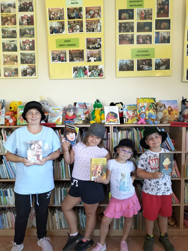 Na tle regałów z książkami i maskotkami stoją dwie dziewczynki, chłopiec i osoba dorosła w kapeluszach i trzymają przed sobą ulubione książki