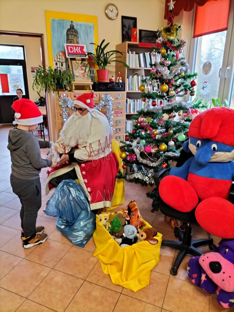 Mikołaj siedzi pod choinką i rozdaje dla dzieci paczki, obok niego jest worek z paczkami i pudełko z maskotkami, w tle regał z książkami, duża maskotka smerfa