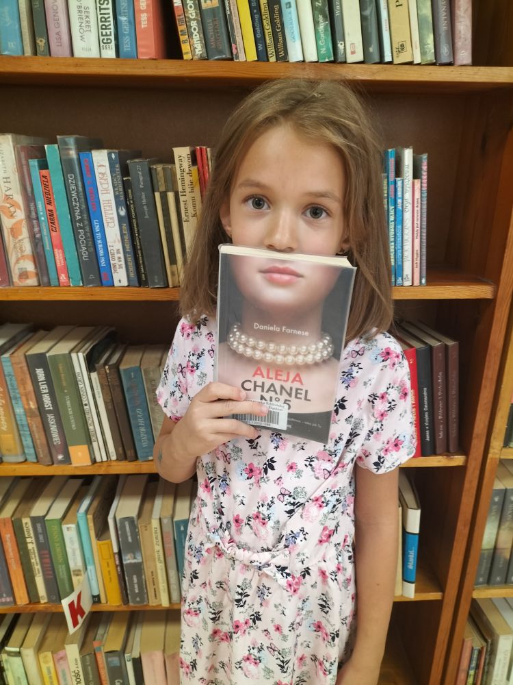 dziewczynka w jasnej sukience, trzyma książkę w ręku i zakrywa tą książką usta, w tle regały z książkami…
