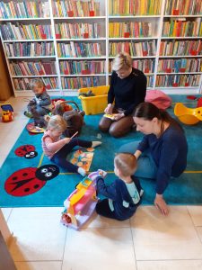 Na zdjęciu widać dzieci siedzące z rodzicami na dywanie i bawiące się zabawkami. W tle regały z książkami.