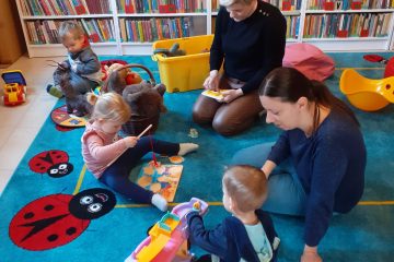 Na zdjęciu widać dzieci siedzące z rodzicami na dywanie i bawiące się zabawkami. W tle regały z książkami.