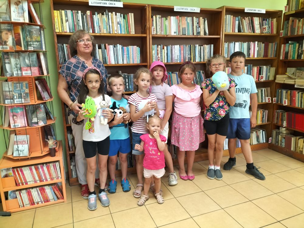 Grupa dzieci wraz panią bibliotekarka stoi na tle regałów z książkami, czworo dzieci trzyma w rękach maskotki.