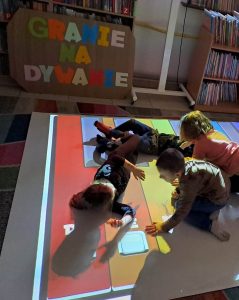 Na zdjęciu widać dzieci na podłodze interaktywnej. W tle kolorowy napis „Granie na dywanie” oraz regały z książkami.