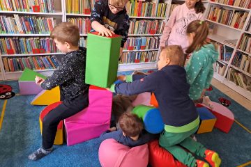 Dzieci wykonują budowlę z dużych, kolorowych, piankowych klocków, w tle regały z książkami.