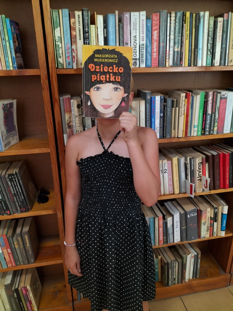 dziewczynka w ciemnej sukience, w ręce trzyma książkę i zakrywa sobie twarz, w tle regały z książkami…