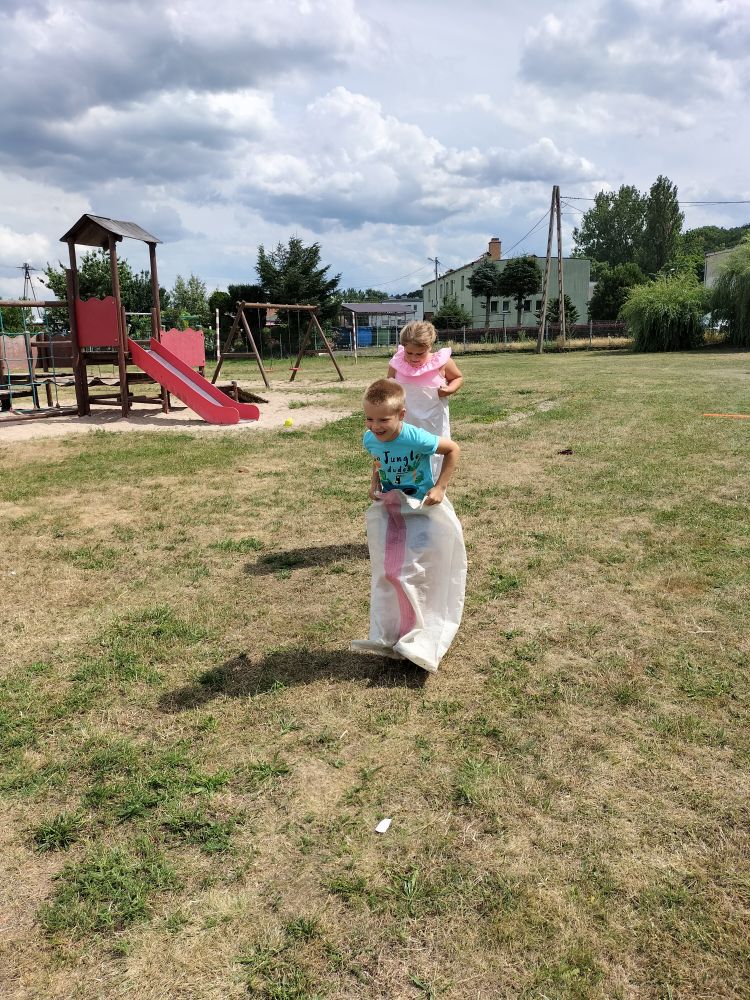 Dziewczynka z chłopcem skaczą w workach na zielonej trawie, w tle widać plac zabaw.