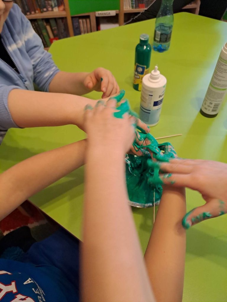 Na zdjęciu widać ręce dzieci oblepione kolorową masą, którą same przygotowały. W tle stół, na którym stoją materiały plastyczne.