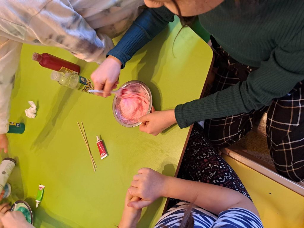 Przy stole siedzą dzieci, dziewczynka miesza w miseczce kolorowa masę. Na stole leżą farby i barwniki spożywcze.