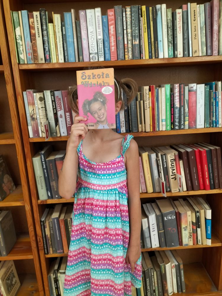 na tle regału z książkami stoi dziewczynka w kolorowej sukience i trzyma książkę, zasłaniając sobie twarz..