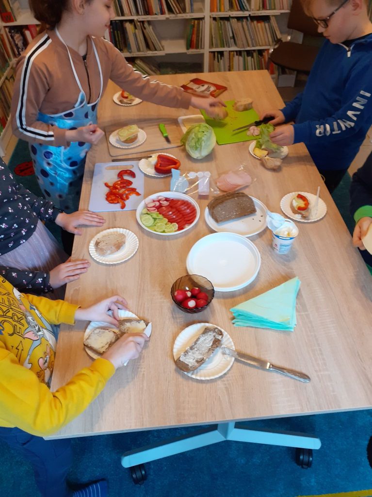 Przy dużym stole stoją dzieci i robią kanapki. Na stole znajdują się : warzywa, masło, wędliny, chleb oraz talerze, noże i deski do krojenia. W tle półki z książkami.