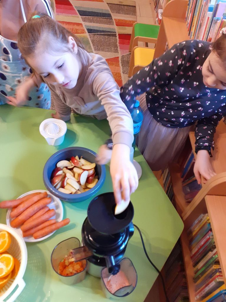 W miskach na stole znajdują się jabłka, pomarańcze i marchewki. Dzieci wrzucają warzywa i owoce do maszyny, która robi soki. W tle dywan i półki z książkami.