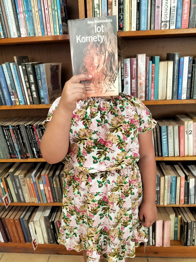 dziewczynka w kolorowej sukience w kwiatki stoi na tle regałów z książkami i w ręce trzyma książkę zakrywając twarz…