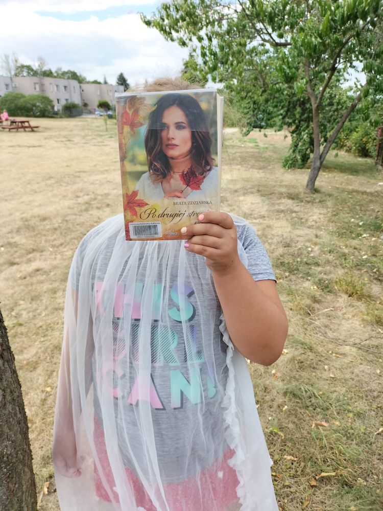 dziewczynka stoi pod drzewem i książką zakrywa sobie twarz, w tle trawa, drzewa i bloki mieszkalne…