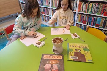 Przy stole siedzą dziewczynki i malują palcami na kartce roztopioną czekoladą, na stoliku znajdują się książki o czekoladzie i rozpuszczona czekolada w miseczce. W tle regały z książkami.