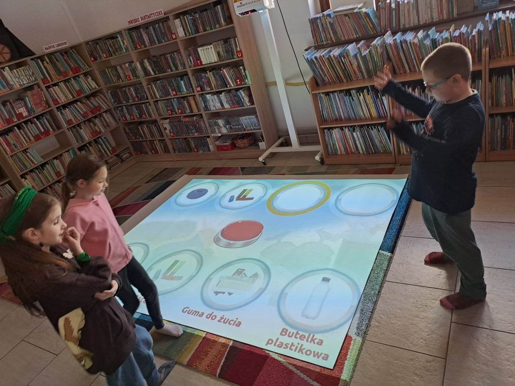 Dzieci stoją przy podłodze interaktywnej, na której wyświetla się gra. W tle półki z książkami.