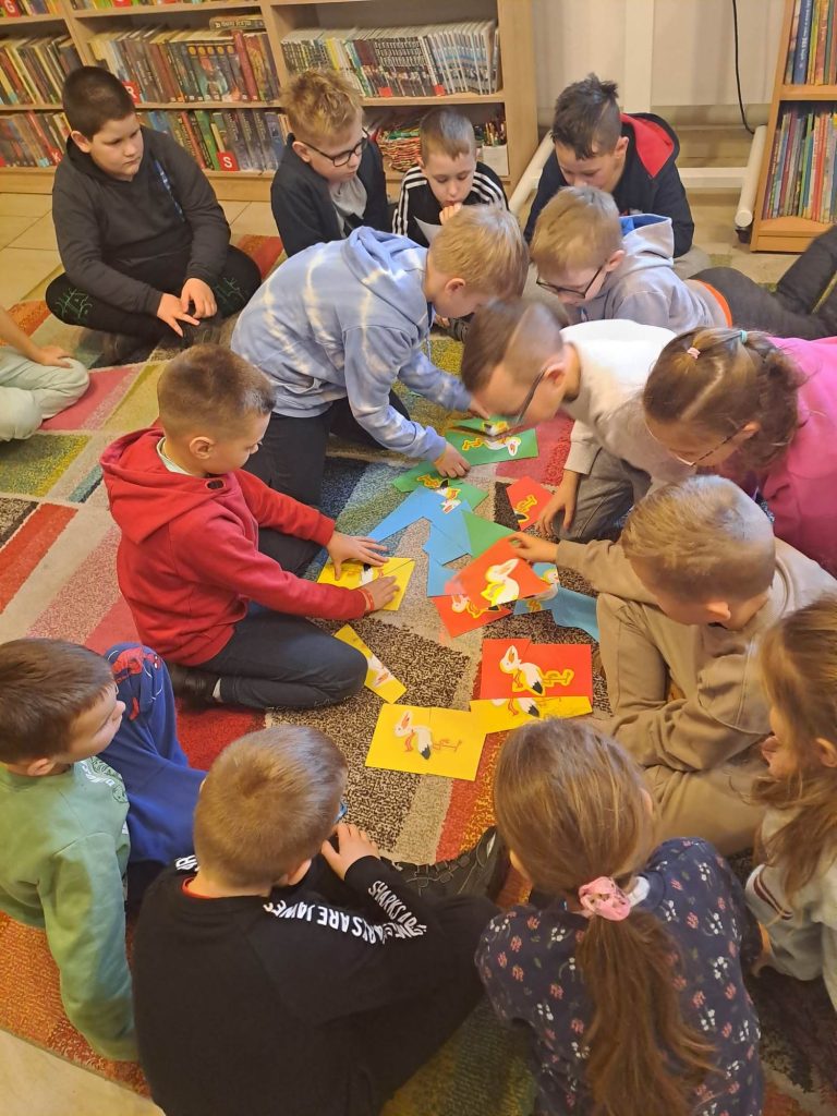 Na dywanie w kręgu kucają dzieci i układają kolorowe wycinanki (puzzle). W tle regały z książkami.
