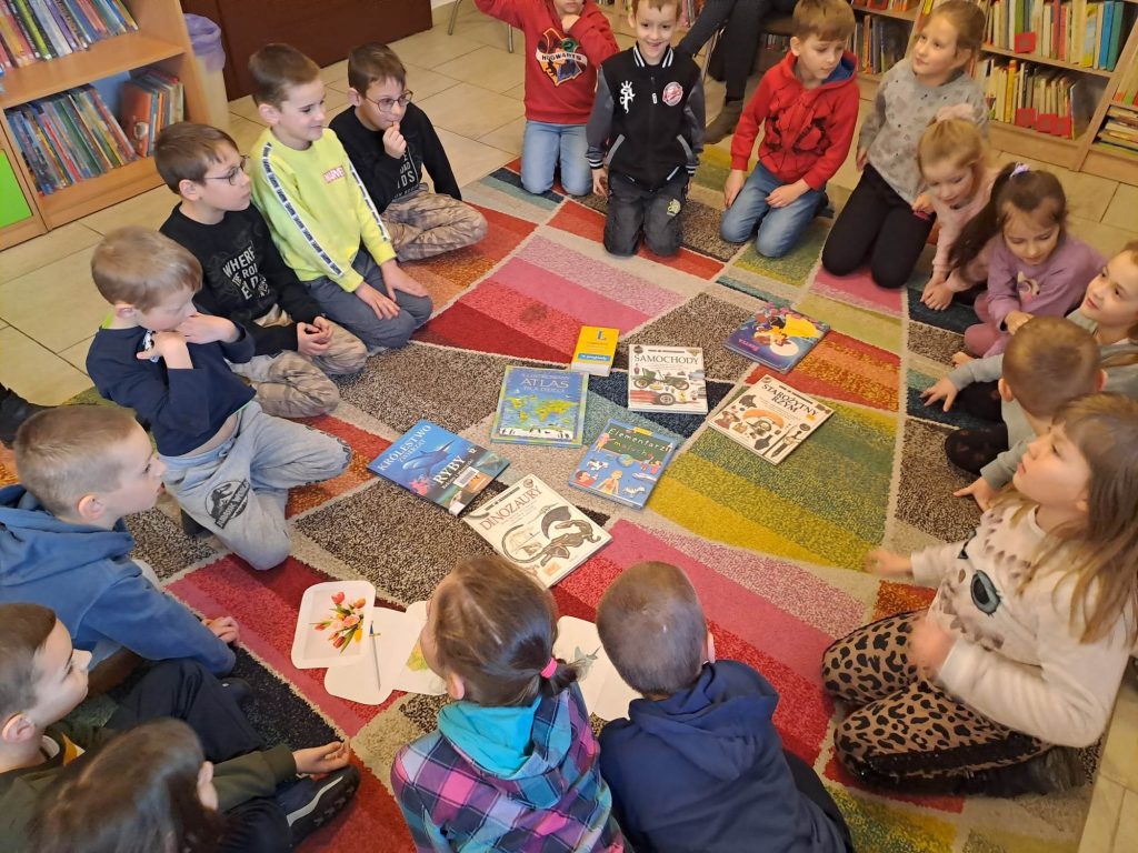 Na dywanie w kółeczku siedzą dzieci, na środku leżą rozłożone różne książki. W tle widać podłogę i regały z książkami.