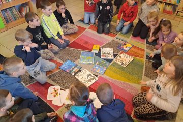 Na dywanie w kółeczku siedzą dzieci, na środku leżą rozłożone różne książki. W tle widać podłogę i regały z książkami.
