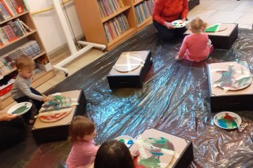 Dzieci siedzą z opiekunami na wyścielonej na dywanie folii. Na podstawkach mają kartonowe jajka i malują je farbami. W tle widać półki z książkami.