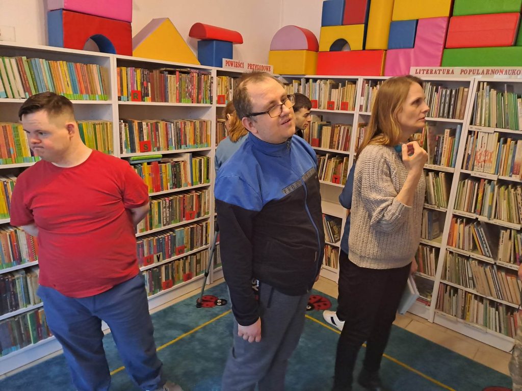 Na zdjęciu widać ludzi, którzy rozglądają się po półkach z książkami. W tle widać regały z książkami, na nich duże , piankowe, kolorowe klocki.