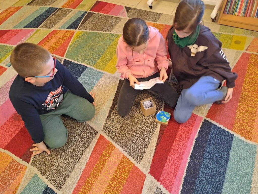 Na dywanie siedzą dzieci. Dziewczynka trzyma w ręku kartkę z której czyta, przed nią stoi otwarte małe pudełeczko z kartonu, w którym widać karteczki.