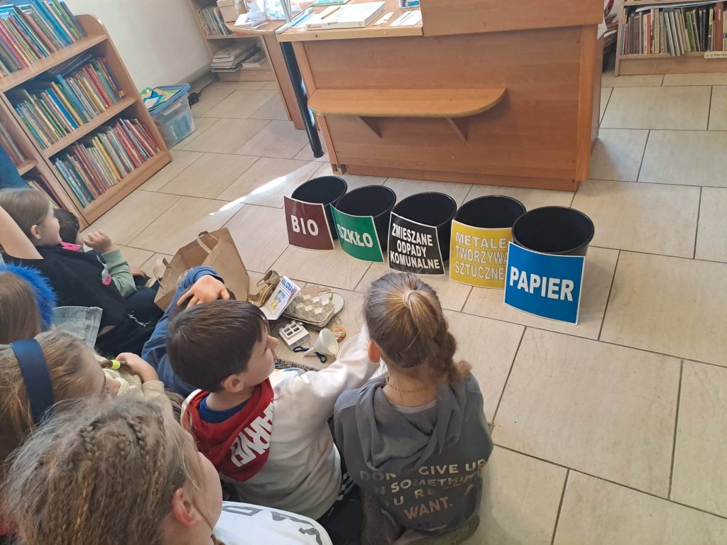 Dzieci siedzą na dywanie, przed nimi leżą odpady i stoją podpisane pojemniki na nie. W tle widać podłogę i książki na półkach.