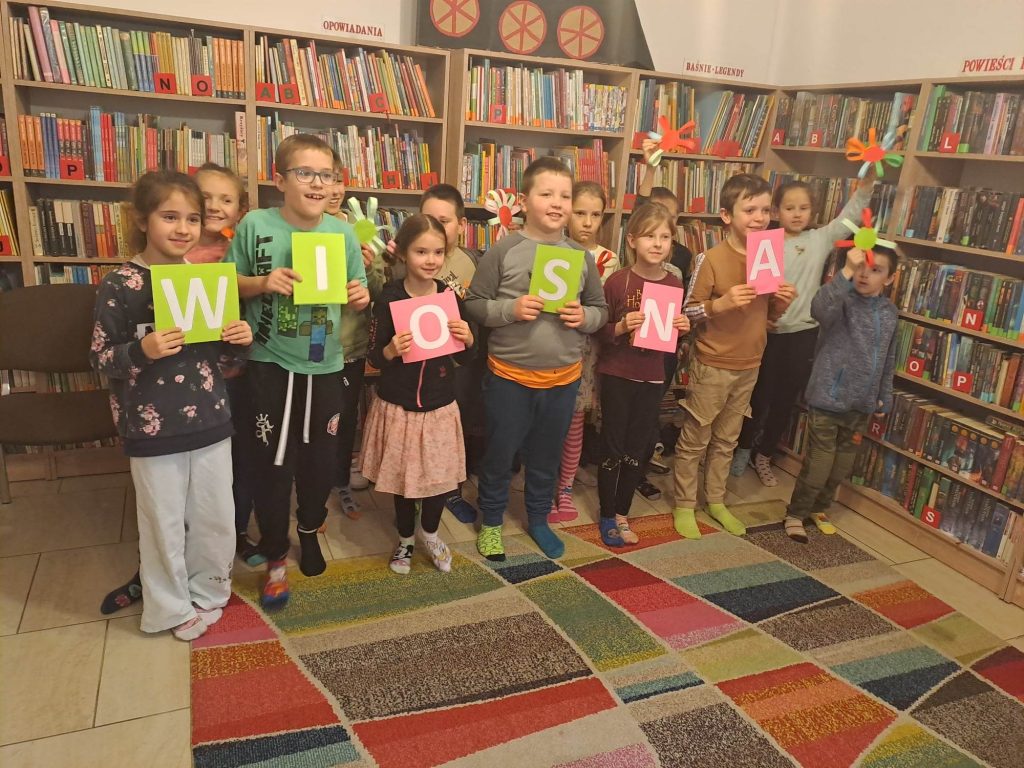 Dzieci stoją w rzędach. W pierwszym rzędzie każde dziecko trzyma w ręku kolorową literę, które razem tworzą napis WIOSNA, a dzieci z tyłu trzymają w górze kolorowe kwiaty. W tle regały z książkami.