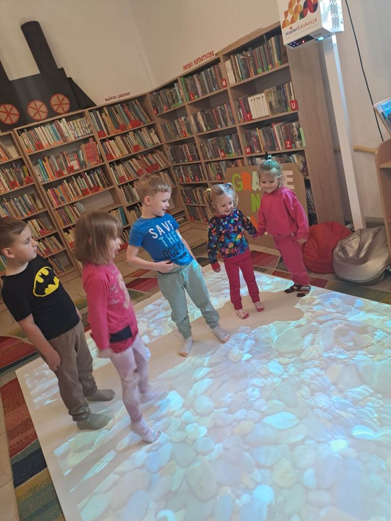 Na podłodze interaktywnej stoją dzieci. W tle widać ściany i regały z książkami.