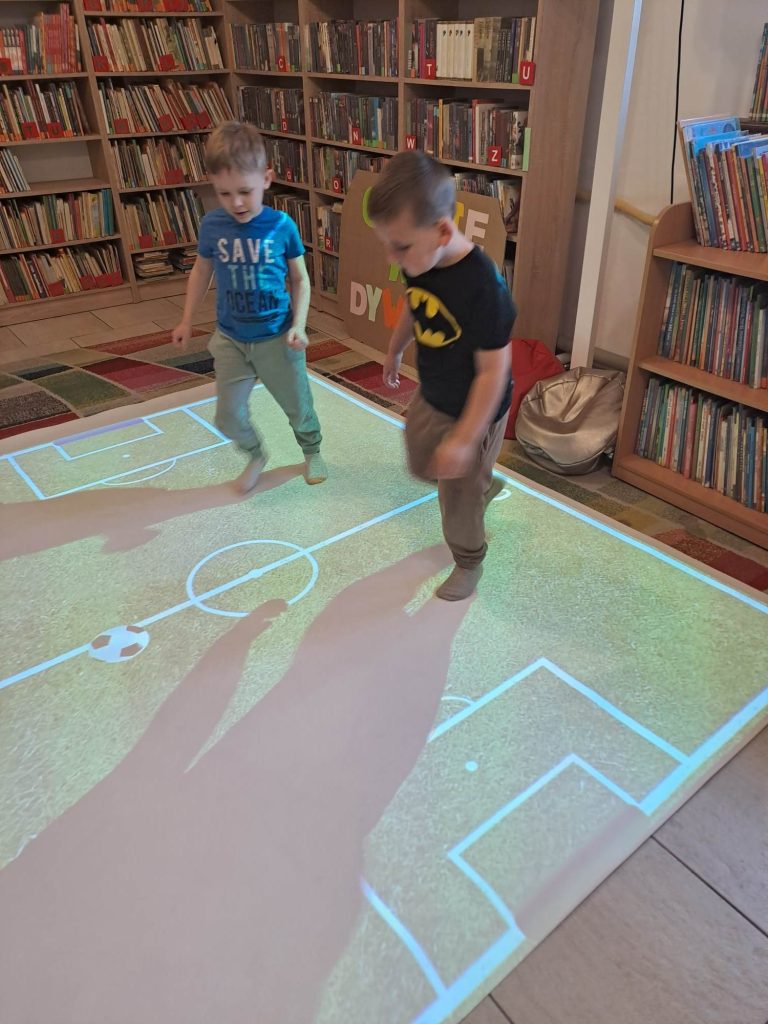 Na podłodze interaktywnej wyświetla się boisko do piłki nożnej i dwaj chłopcy grają. W tle regały z książkami.