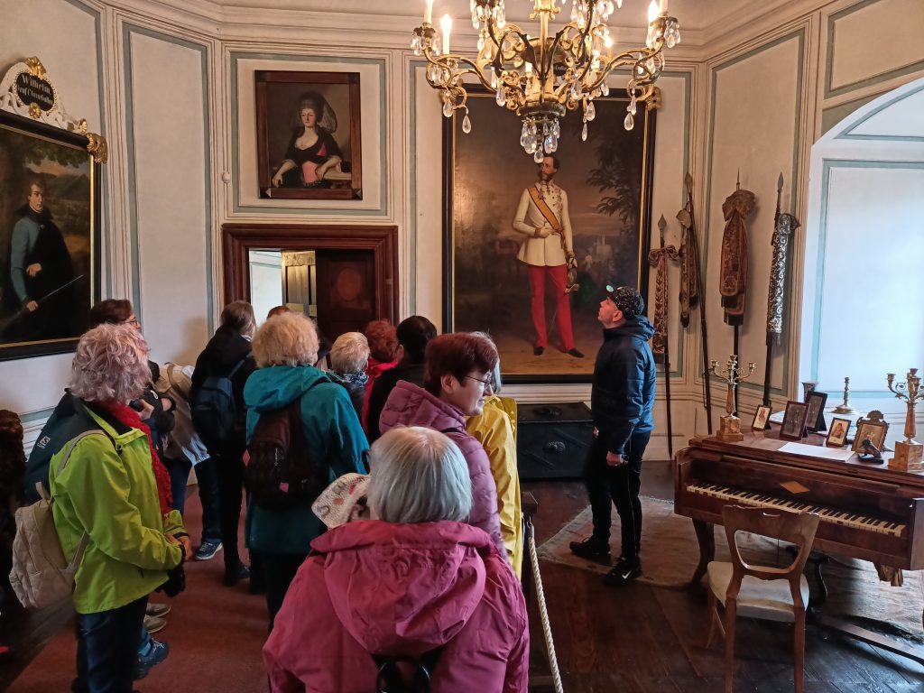 Z lewej strony grupa osób stojących tyłem. Z prawej strony fortepian. W tle obrazy na ścianach i duży żyrandol.
