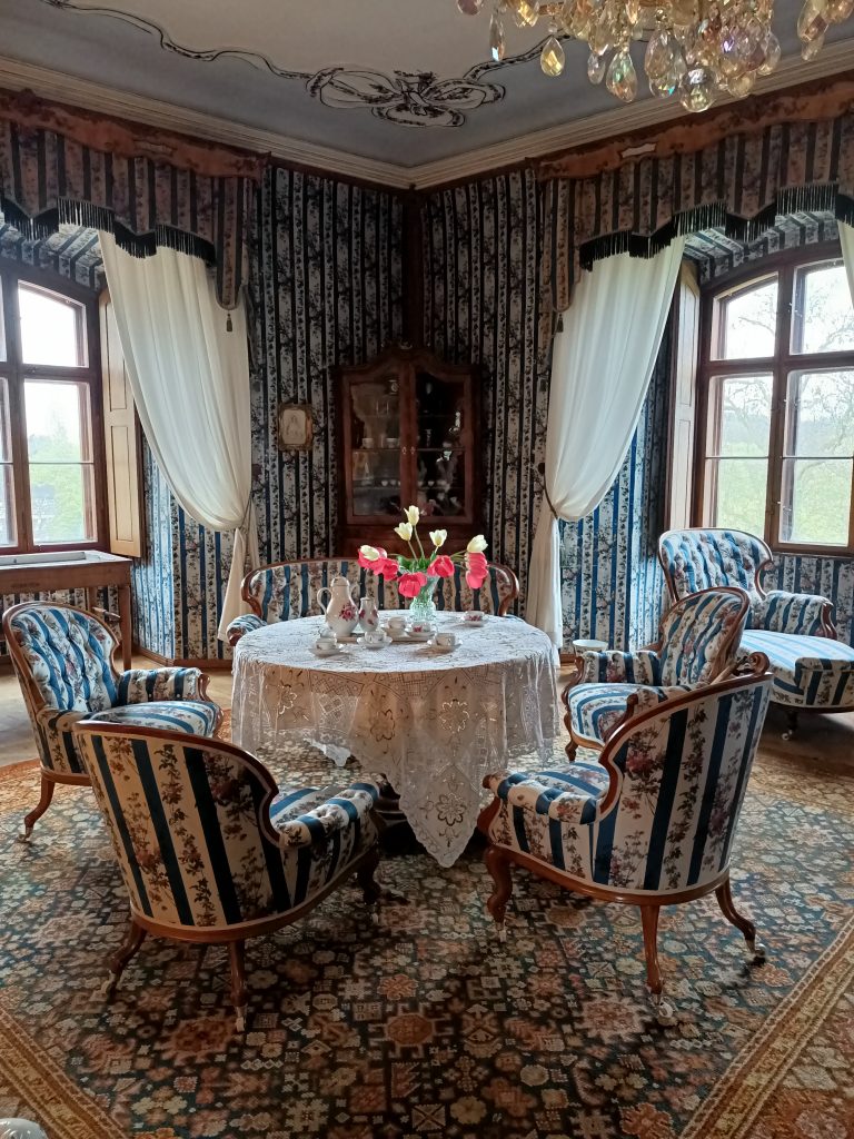Pokój pałacowy. Na środku okrągły stolik, na nim filiżanki, dzbanek, wazon z kwiatami. Wokół fotele. W tle, z lewej i prawej strony, dwa duże okna. Pośrodku oszklony kredens.