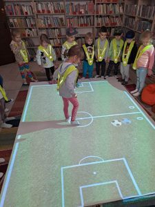 Na podłodze interaktywnej wyświetla się boisko do piłki nożnej i na nim znajduje się dziewczynka, dookoła stoją dzieci. W tle widać regały z książkami.