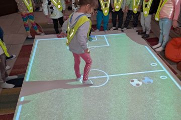 Na podłodze interaktywnej wyświetla się boisko do piłki nożnej i na nim znajduje się dziewczynka, dookoła stoją dzieci. W tle widać regały z książkami.