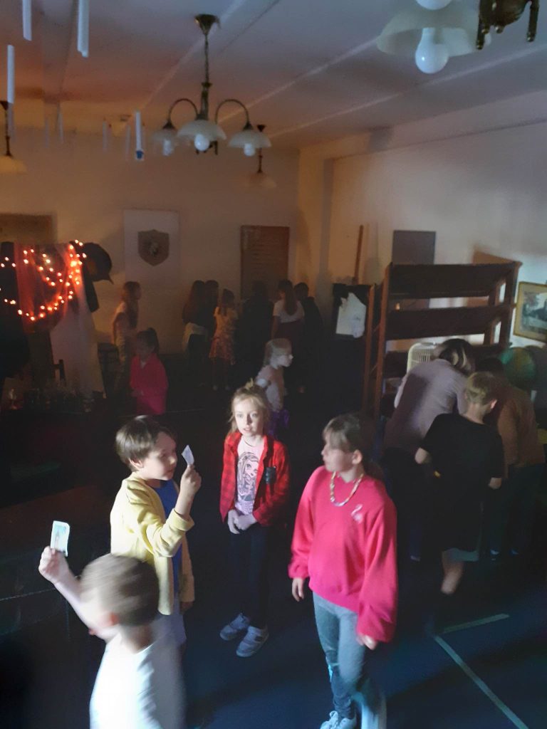 Na zdjęciu widać dzieci, które znajdują się w pomieszczeniu o przygaszonym świetle dwoje trzyma w ręku karty, w tle ściany, żyrandole, półki i obrazy.