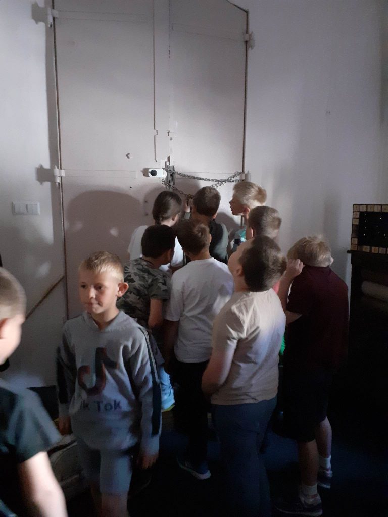 Na zdjęciu znajdują się dzieci, próbują otworzyć łańcuch z kłódką na który zamknięte są drzwi, w tle ściana i drzwi.