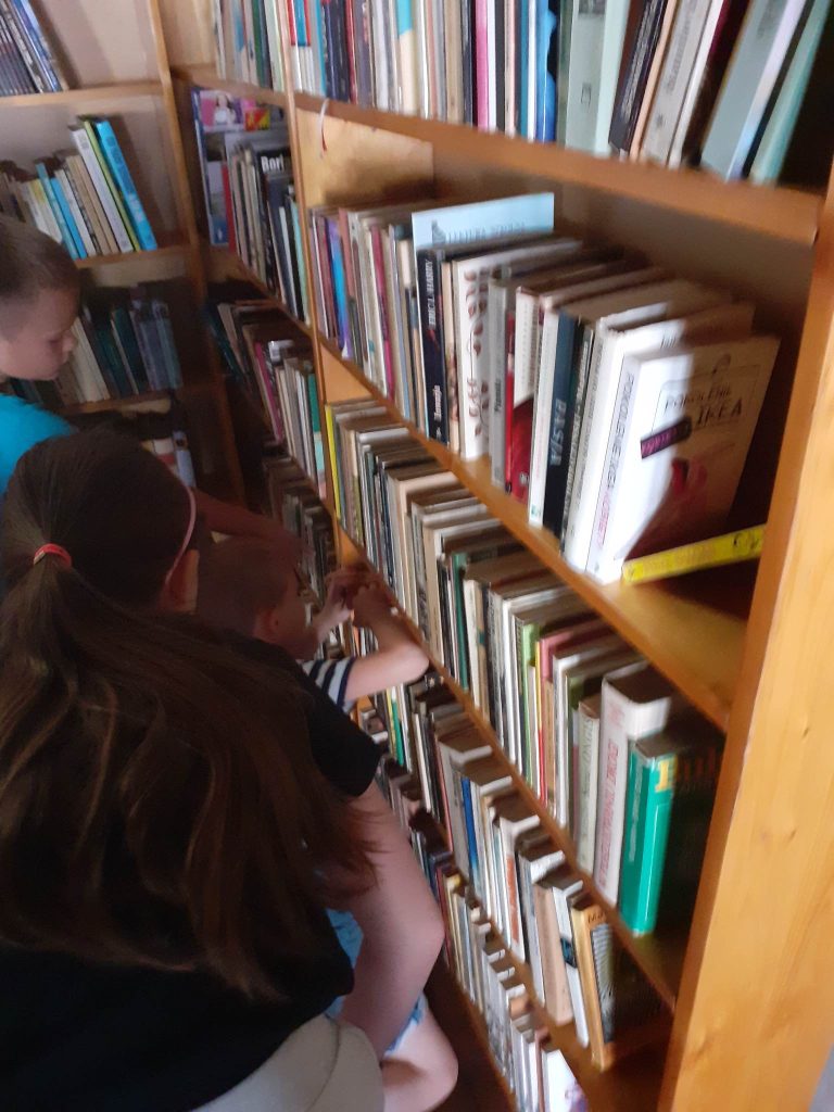 Na zdjęciu dzieci stoją przed regałami z książkami. Jedna z tych osób przekłada książki na półce.