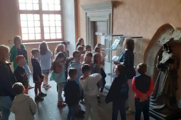Na zdjęciu stoi grupka dzieci wraz z opiekunami, słuchają pani - pracownika Placówki historyczno-muzealnej, która stoi przed nimi. Obok znajduje się szklana gablota z towarem, a w tle jest duże okno.