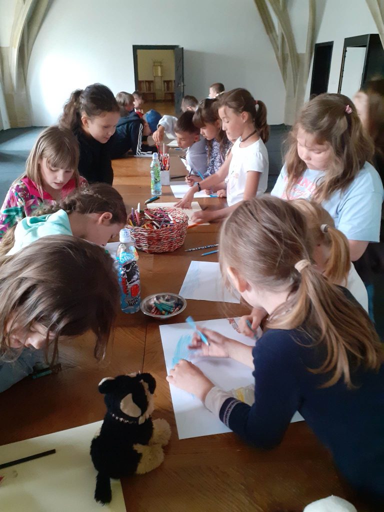 Grupka dzieci stoi wkoło długiego stołu na, którym leżą kredki, kartki. Dzieci malują kolorowe obrazki.