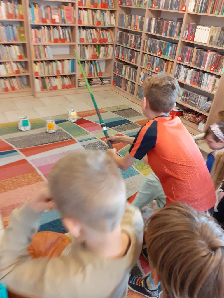 Na kolorowym dywanie znajdują się trzy koszyki. Jedna osoba z grupki dzieci trzyma wędkę. W tle regały z książkami.