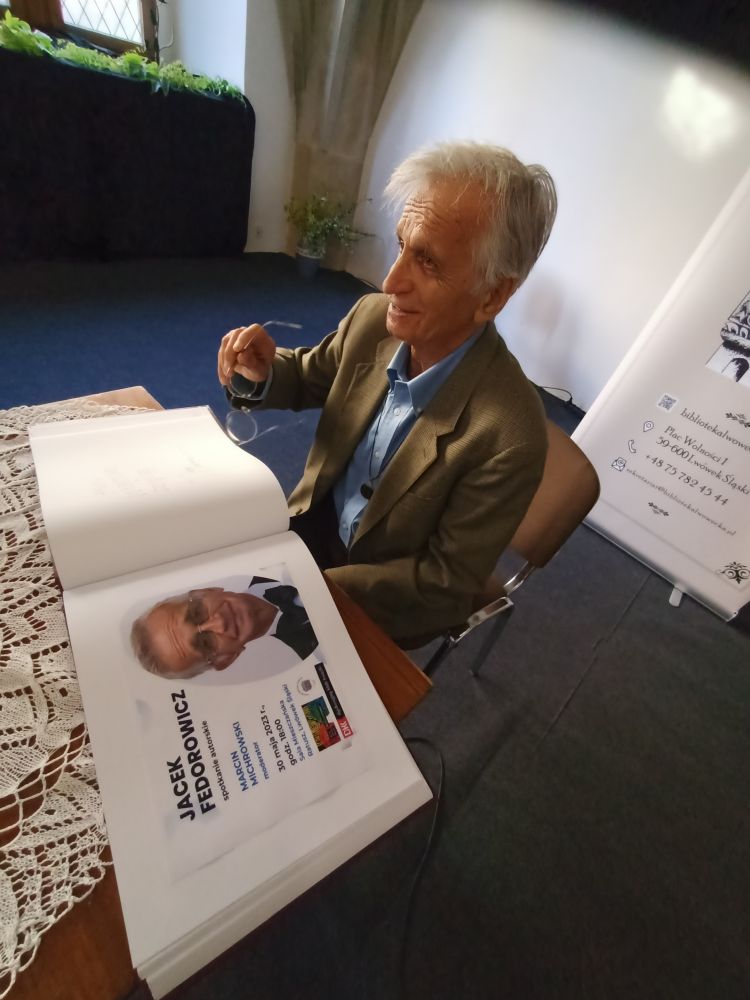 Mężczyzna siedzący bokiem przy stoliku, unoszący w prawej dłoni okulary. Przed nim, na stoliku, otwarta księga z plakatem, na którym widnieje portret mężczyzny i napis „Jacek Fedorowicz”.