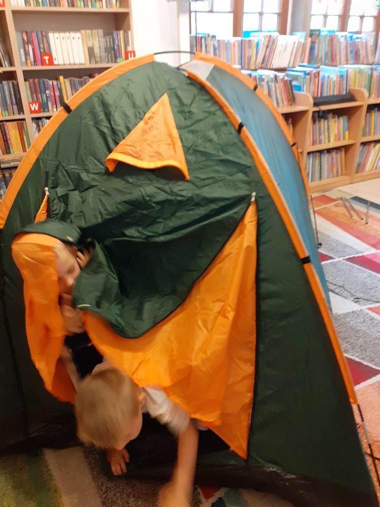Na środku sali rozłożony jest namiot, z którego wychodzi dwóch chłopców. W tle regały z książkami i okna.