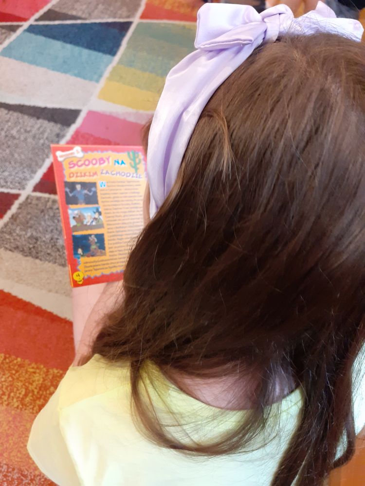 Na kolorowym dywanie siedzi tyłem dziewczynka. W rękach trzyma książkę.