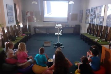 W sali stoi duży ekran i projektor. Dzieci siedzą na kolorowych pufach i mają miski z popcornem.